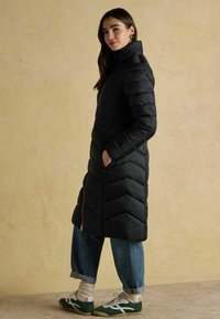 Garšs melns pūkains mētelis ar chevrona kvadrātveida rakstu, augstu apkakli, aizdari ar rāvējslēdzēju, apvienots ar galiem uzkāptiem zilo džinsu un zaļām kedām.