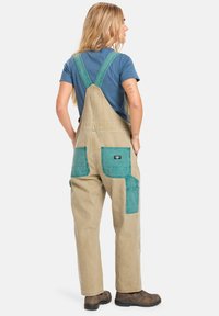 Salopette beige con tasche turchesi, dotate di spalline regolabili e una vestibilità rilassata. Il materiale sembra resistente e ha una consistenza morbida. Scarpe stivali marroni indossate.
