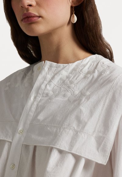 White cotton shirt with a high collar and floral embroidery detail. Features a button closure and draped fit, paired with shell earrings.