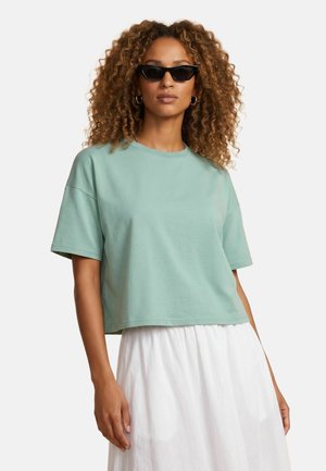 Frau mit lockigem Haar trägt schwarze Sonnenbrille, ein lockeres mintgrünes T-Shirt und einen weißen Rock und steht vor einem schlichten Hintergrund.