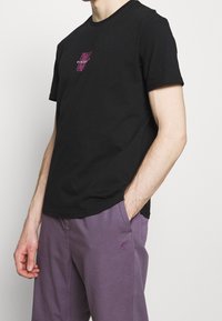 T-shirt en coton noir à manches courtes et col rond, orné d'un graphique coloré "DISCOUNT" sur la poitrine. Associé à un pantalon violet.
