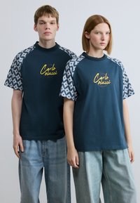 Μπλε ναυτικά t-shirts με κοντά μανίκια σε σχέδιο, με κίτρινο λογότυπο "Carlo Colucci". Χαλαρή εφαρμογή, συνδυασμένα με ανοιχτόχρωμο τζιν παντελόνι.