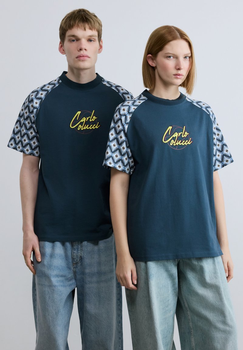 Μπλε ναυτικά t-shirts με κοντά μανίκια σε σχέδιο, με κίτρινο λογότυπο "Carlo Colucci". Χαλαρή εφαρμογή, συνδυασμένα με ανοιχτόχρωμο τζιν παντελόνι.