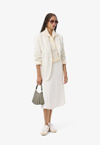 Donna con i capelli intrecciati che indossa occhiali da sole, un blazer crema, un maglione con colletto, una gonna plissettata e sneakers bianche, tenendo in mano una piccola borsa grigia.