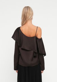 Top marrone senza spalle con un design a strati di volant, maniche lunghe e spalline regolabili, abbinato a una gonna nera plissettata.