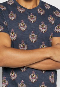 Mörkblå t-shirt med ett upprepande paisleymönster i rött, gult och vitt. Rund halsringning och korta ärmar med en mjuk textur.