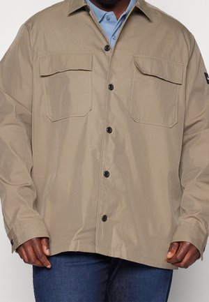 Homme portant une veste beige à boutons avec deux poches poitrine sur une chemise bleue à col et un jean bleu foncé, visible du cou aux hanches.