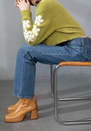 Personne assise sur une chaise portant un pull vert duveteux avec des motifs de fleurs blanches, un jean bleu et des bottines beiges à talons.