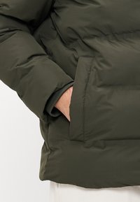 Person's hand inside olive green puffer jacket pocket, showing jacket's padded texture and cuff detail over light-colored pants.