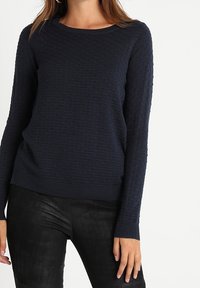 Granatowy sweter z dzianiny o teksturowanym wzorze, okrągłym dekolcie, długich rękawach i ściągaczu u dołu. Połączony z czarnymi skórzanymi spodniami.