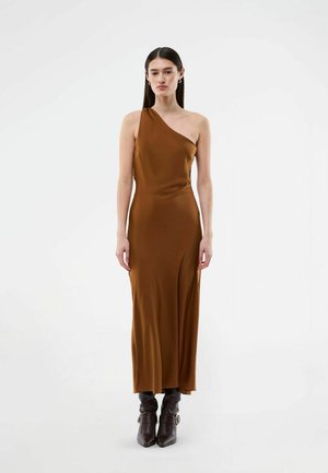 Kvinde iført en brun satin kjole med ét skulderstykke og mørkebrune læderstøvler med spids tå, stående mod en ensfarvet hvid baggrund.