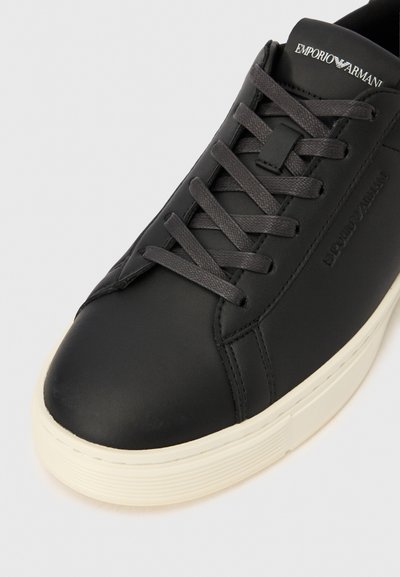 Zapatilla deportiva de cuero negro con cordones planos, logo en relieve y suela de goma color crema. Presenta un diseño elegante, minimalista y un panel lateral con textura.