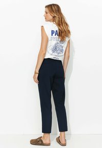 T-shirt blanche sans manches avec un design graphique bleu et du texte au dos, associée à un pantalon marine et des chaussures à enfiler marron. Cheveux longs visibles.