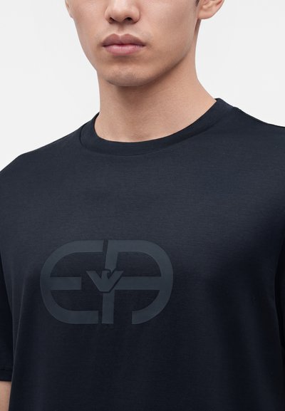 Νέος άνδρας φοράει μαύρο T-shirt με στρογγυλή λαιμόκοψη και σκούρο ανάγλυφο γεωμετρικό λογότυπο στο στήθος, σε απλό φόντο.