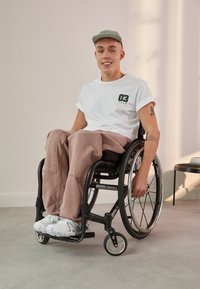 Silla de ruedas negra con llantas de aleación, asiento acolchado y reposapies. Modelo vistiendo una camiseta blanca, pantalones rosa claro y zapatos blancos, sentado en un piso gris.