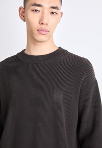 Maglione a coste di un marrone scuro, con scollo rotondo e un delicato patch logato in rilievo sul petto. Tessuto morbido e vestibilità rilassata.