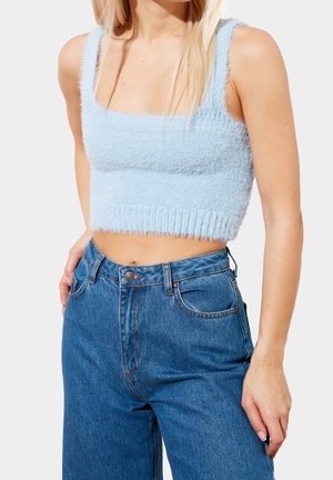 Frau trägt ein hellblaues, flauschiges, kurzes Tanktop kombiniert mit hoch taillierter blauer Jeans, Hand auf der Hüfte ruhend.