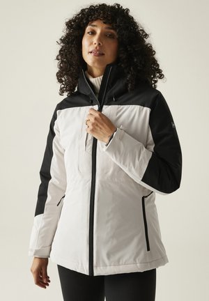 Femme aux cheveux bouclés portant une veste zippée blanche et noire avec un col montant et des poches latérales à fermeture éclair, ajustant la fermeture éclair.