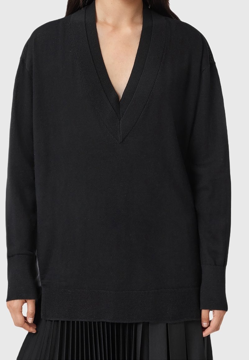Femme portant un pull noir à manches longues avec col en V associé à une jupe plissée noire sur un fond uni.