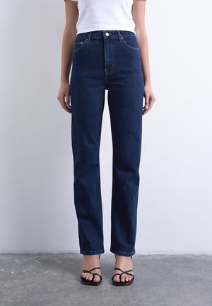 Person iført mørkeblå højtaljede jeans med lige ben, knap og lynlås, kombineret med sorte sandaler med stropper og en hvid top.