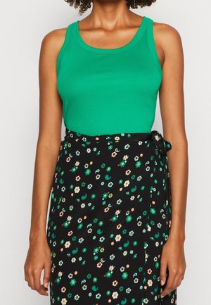 Frau trägt ärmelloses grünes Oberteil und schwarzen, hoch taillierten Rock mit kleinem grün-weißem Blumenmuster und seitlicher Schleife.