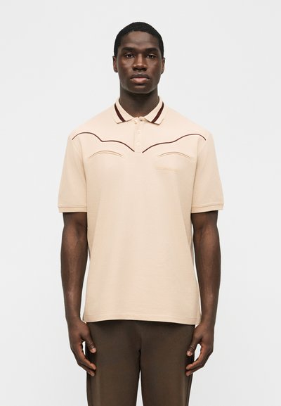 Άνδρας που φοράει μπεζ πόλο με κοντά μανίκια και καφέ λεπτομέρεια στο τελείωμα, καθώς και σκούρο καφέ παντελόνι, στέκεται μπροστά σε απλό ανοιχτό φόντο.