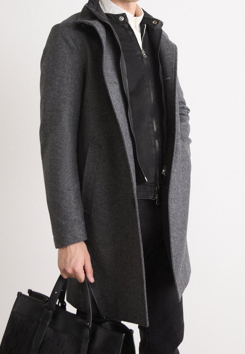 Personne vêtue d'une tenue superposée avec un manteau en laine gris et une veste noire zippée tenant un sac fourre-tout en cuir noir par les poignées.