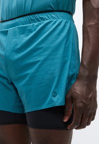 Τυρκουάζ αθλητικό σορτς από ελαφρύ ύφασμα με ελαστική μέση και κρυφό τσέπη. Φορεμένο πάνω από μαύρο σορτς συμπίεσης.