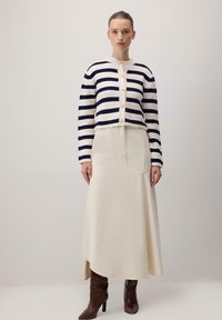 Prugasti kardigan u krem i tamnoplavoj boji s zlatnim dugmadima, uparen s krem suknjom i smešnim čizmama do koljena, s naglašenim krojem.