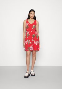 Vestido floral vermelho com decote arredondado, cintura ajustada e barra rodada. Apresenta flores cor-de-rosa e folhas verdes, usado com sandálias brancas de plataforma.