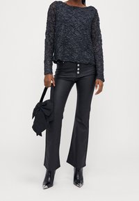 Schwarzes, transparentes Langarmoberteil mit strukturiertem Muster; eng anliegende schwarze Hose mit drei Knöpfen; schwarze Handtasche mit Schleifendetail; spitze Stiefel.