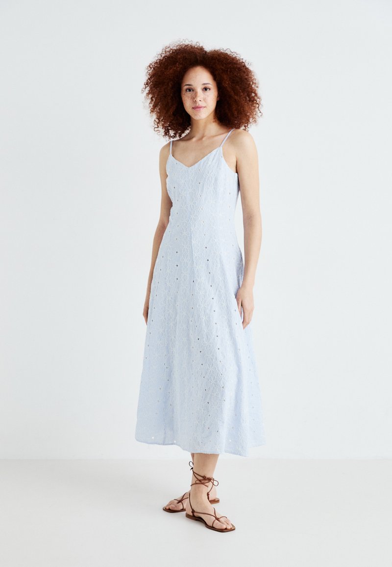 Rochie midi de culoare albastră deschisă cu bretele subțiri, având modele perforate și o siluetă fluidă. Purtați cu sandale maro cu șiret.