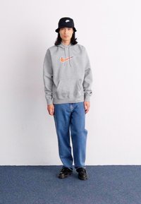En person står foran en ensfarvet hvid væg, iført en grå Nike hættetrøje, blå jeans og sorte sko, med en sort bøttehat.