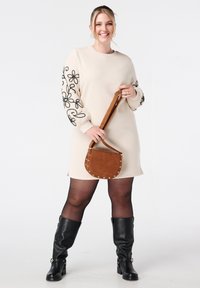 Beige jurk met zwarte bloemenborduursels op de mouwen, gecombineerd met een bruine, gestructureerde ronde tas en kniehoge zwarte leren laarzen.