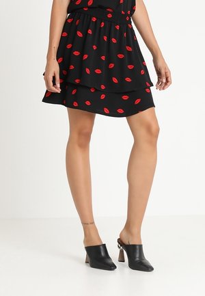 Kobieta ubrana w czarną, warstwową spódnicę z czerwonymi nadrukami ust oraz czarne sandały na obcasie, stojąca na tle jednolitej białej ściany.