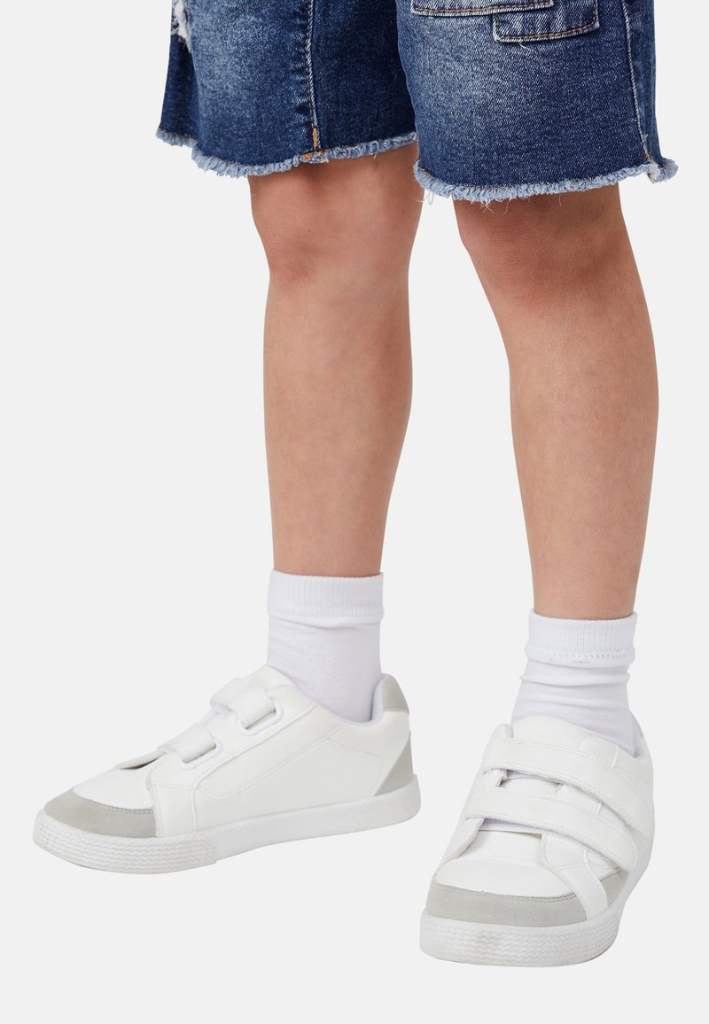 Baskets blanches avec des accents gris, dotées de deux bandes Velcro ajustables, de semelles texturées et d'un dessus lisse. Portées avec des chaussettes blanches.