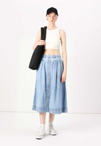 Kevyt denim-midi-hame, jossa on edessä napit ja rypistetty vyötärö, yhdistettynä tiukkaan valkoiseen toppiin ja mustaan lippalakkiin, kantaa mustaa laukku.