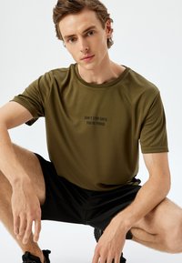 Camiseta atlética de manga corta en verde oliva con cuello redondo y texto negro "NO TE DETENGAS HASTA QUE ESTÉS ORGULLOSO" centrado en la parte delantera.