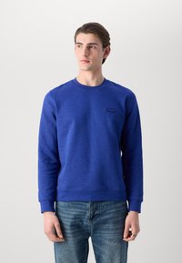 Felpa blu royal con scollo tondo, polsini e orlo a coste. Presenta un piccolo etichetta sul petto. Abbinata a jeans in denim chiaro.