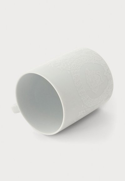 Mug en céramique blanche avec un subtil motif floral et cœur en relief, positionné sur le côté avec la anse visible sur la gauche.