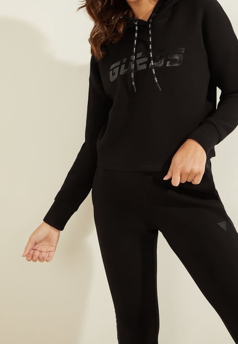 Sweat-shirt à capuche noir avec un logo "GUESS" brillant, accompagné d'un pantalon noir fuselé avec un détail de logo triangulaire sur la jambe.