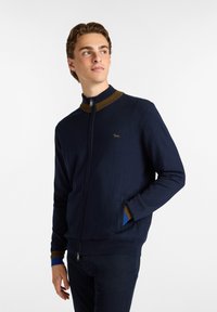 Sudadera navy con cremallera y cuello alto, con acentos marrones y azules en el cuello y los puños; textura suave con detalle de logo.