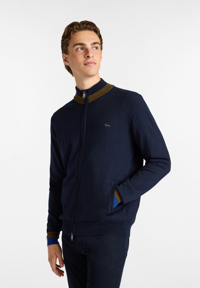 Sudadera navy con cremallera y cuello alto, con acentos marrones y azules en el cuello y los puños; textura suave con detalle de logo.