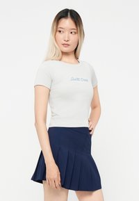 Világosszürke, rövid ujjú, testhez álló póló, amelyen kék színnel hímezve szerepel a "Santa Cruz" felirat. Navy kék, pliszírozott szoknyával párosítva. Egyszerű dizájn, minta nélkül.