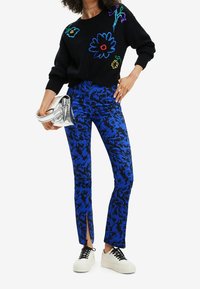 Schwarzer Strickpullover mit mehrfarbigem Blumenstickereien, kombiniert mit blauen gemusterten Hosen mit Schlitzen am Saum und weißen Sneakers. Silberne Clutch.