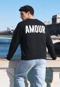 Jeune homme en sweat-shirt noir avec le texte « AMOUR » appuyé sur une balustrade en pierre au bord de la rivière, paysage urbain et pont en arrière-plan sous un ciel clair.