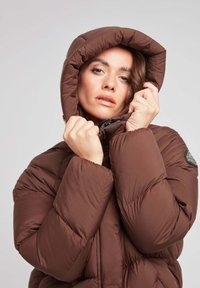 Brązowa kurtka puchowa z kapturem, z pikowanym wzorem, zamkiem błyskawicznym i naszywką z logo. Materiał wydaje się gładki i ocieplany.