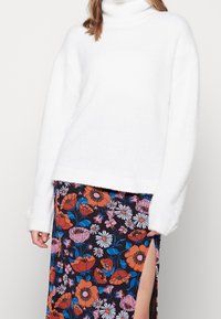 Un maglione a collo alto bianco e soffice con maniche lunghe, abbinato a una gonna floreale con grandi fiori arancioni e blu su uno sfondo scuro.