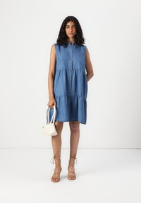 Τζιν φόρεμα χωρίς μανίκια με κουμπιά μπροστά, μαζεμένη μέση, ανοιχτό μπλε χρώμα και άνετη εφαρμογή. Συνδυασμένο με μια μικρή λευκή τσάντα και σανδάλια με λουράκια.