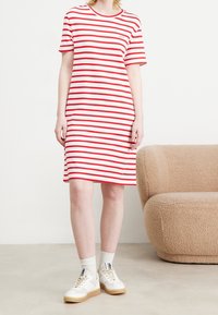 Femme portant une robe blanche longueur genoux avec des rayures horizontales rouges, des chaussettes blanches et des baskets blanches, se tenant près d'un canapé beige texturé.